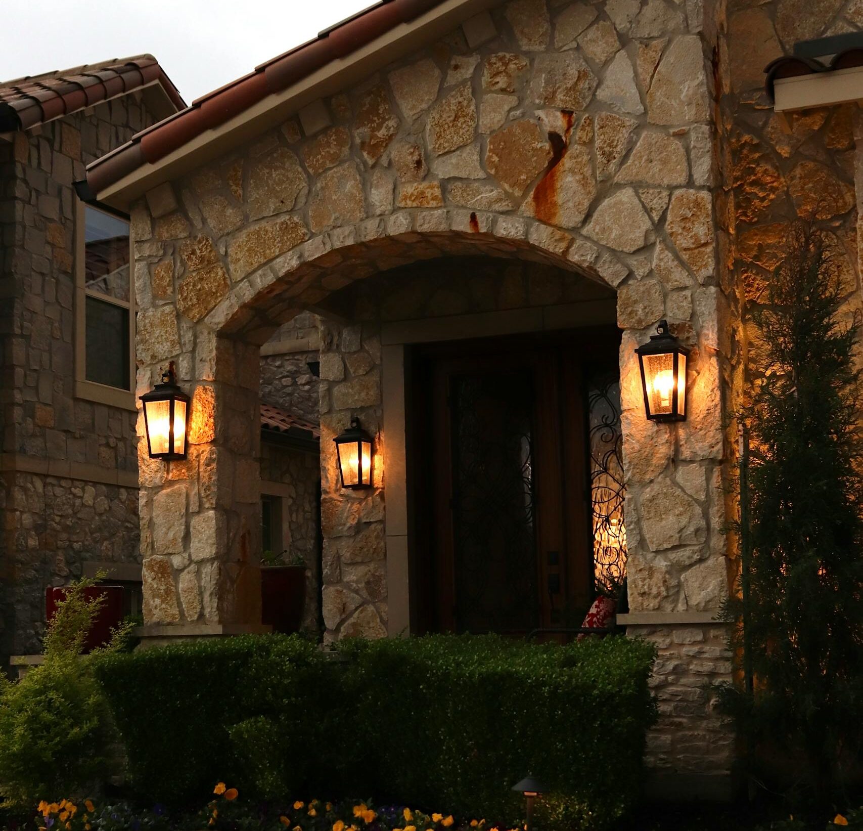 Charming stone entrance illuminated by classic lamps, surrounded by manicured greenery.